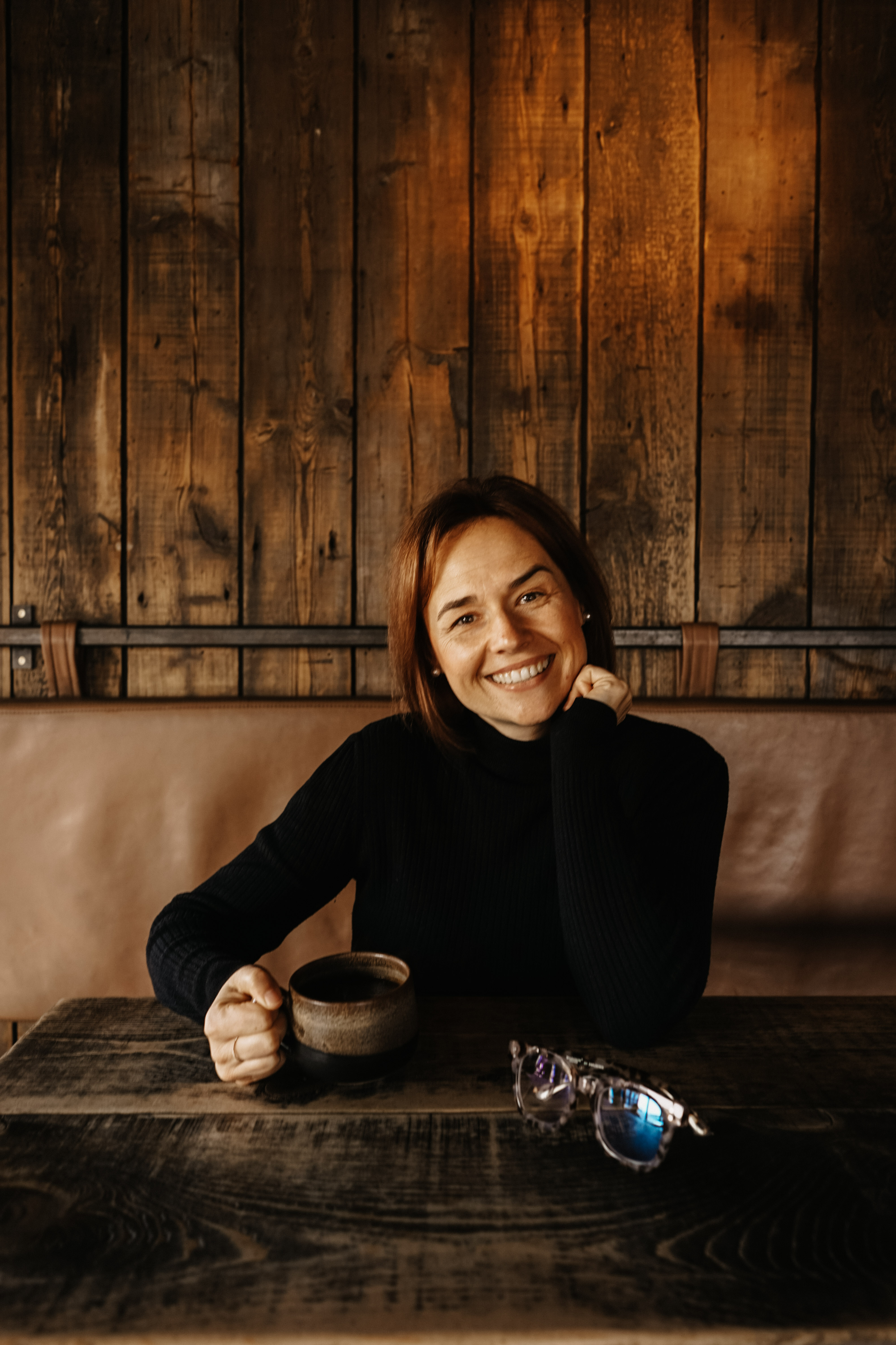 A woman in a black jumper smiling, sitting at a wooden table with a mug of coffee.